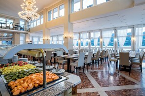 Uma área de buffet com diversas frutas e legumes, várias mesas de jantar com cadeiras e um candelabro.