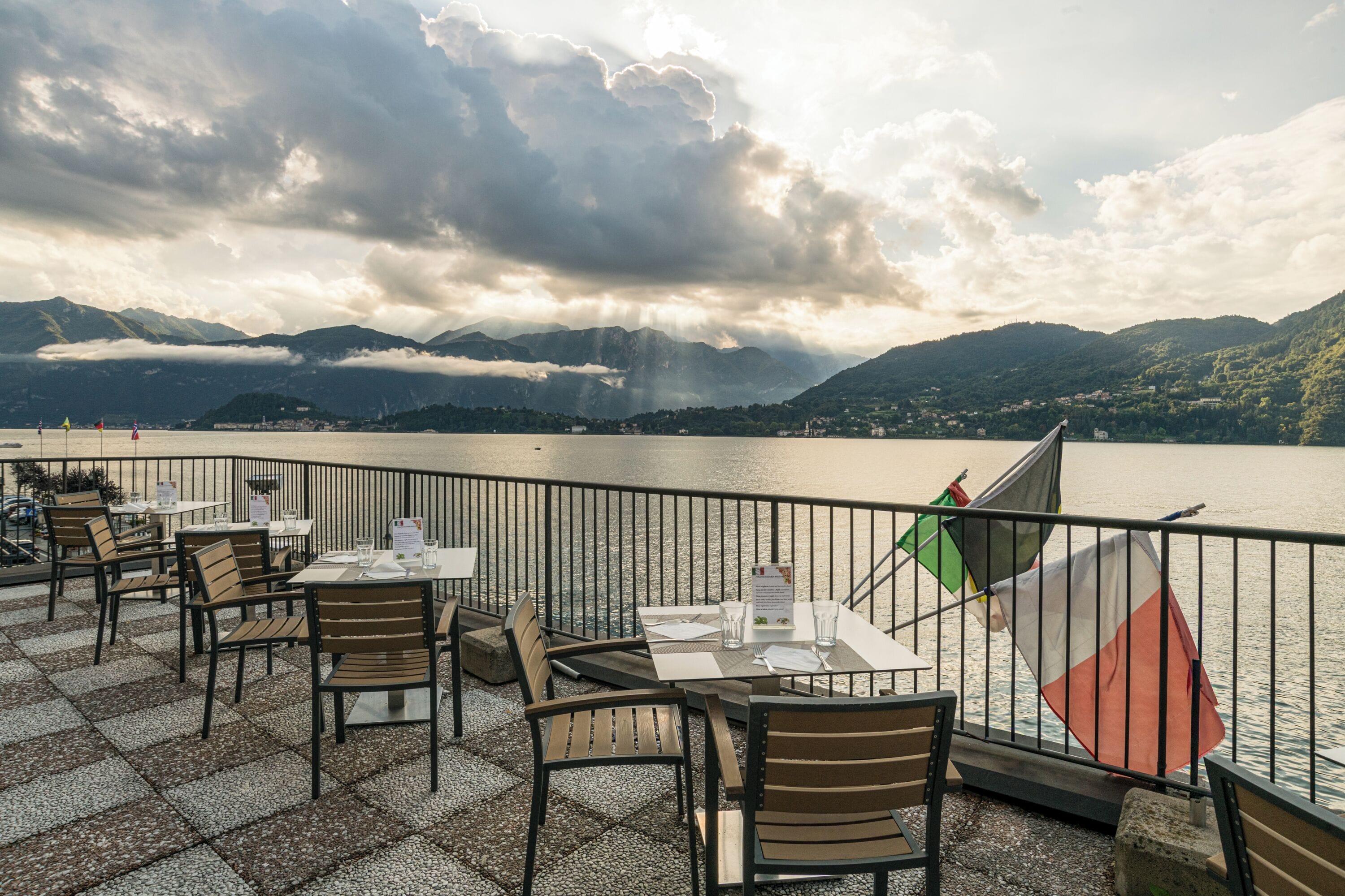 Un área de comedor al aire libre con mesas y sillas, con vista a un lago y montañas.