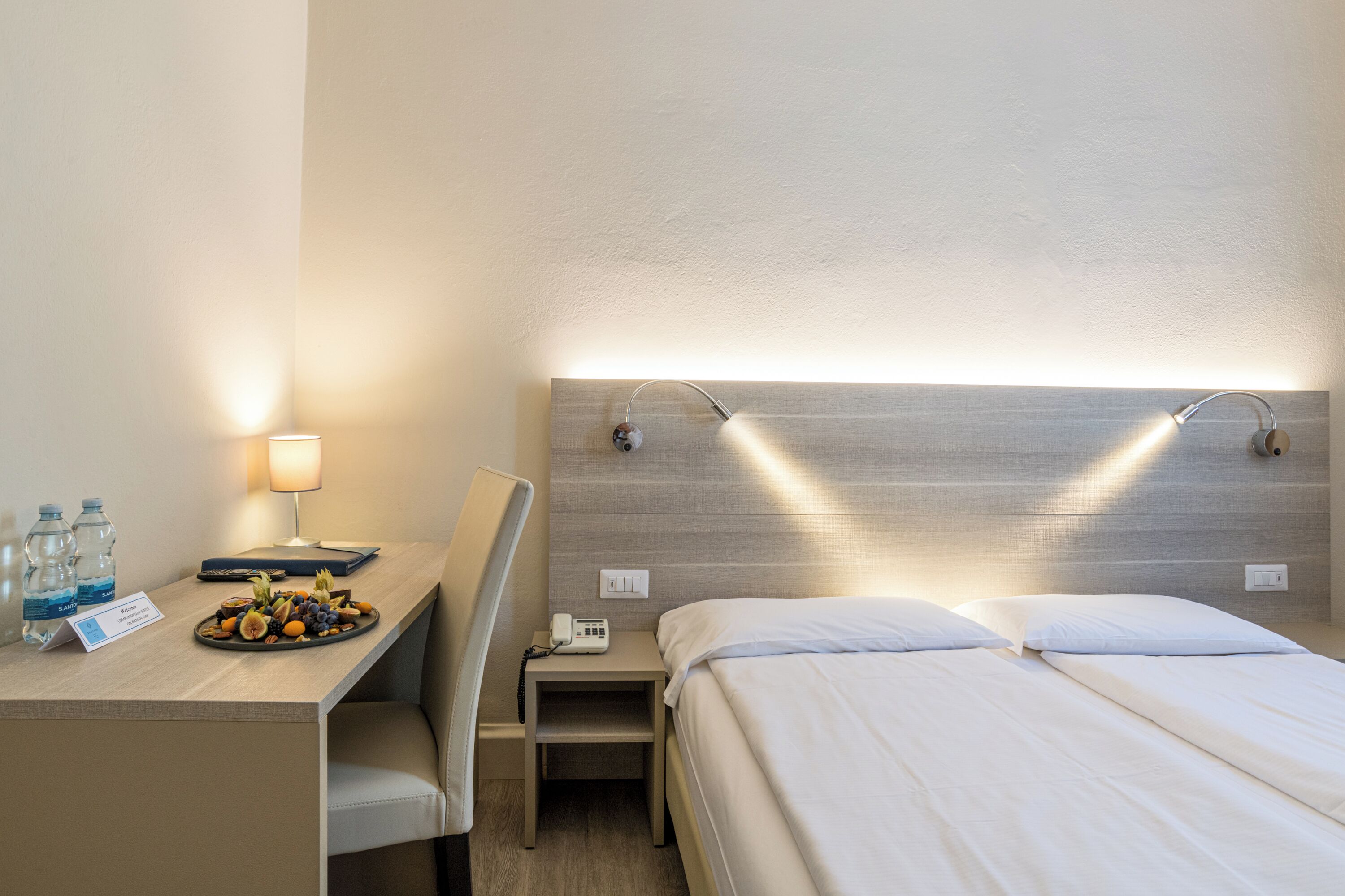 Ein Hotelzimmer mit Bett, Nachttisch, Lampe und einem kleinen Schreibtisch mit Obstplatte und Wasserflaschen.