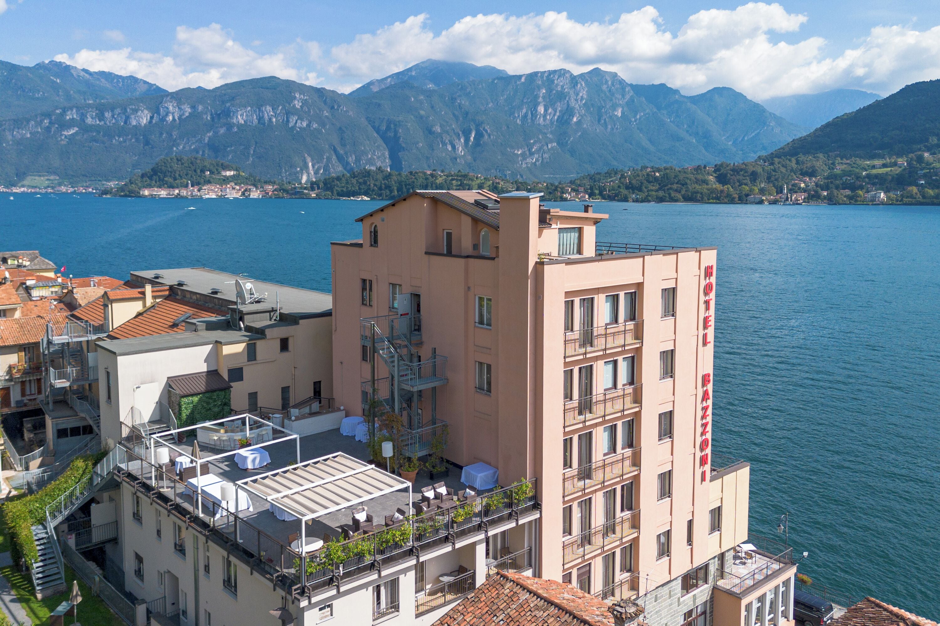 A hotel with a rooftop terrace, a view of the lake, and mountains in the background.