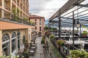 Uma área de refeições ao ar livre coberta, com mesas e cadeiras, vista para um edifício com varandas e um canal ao fundo.