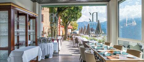 A restaurant with a long table set for dining, a glass display case with wine glasses, and a view of mountains through large windows.