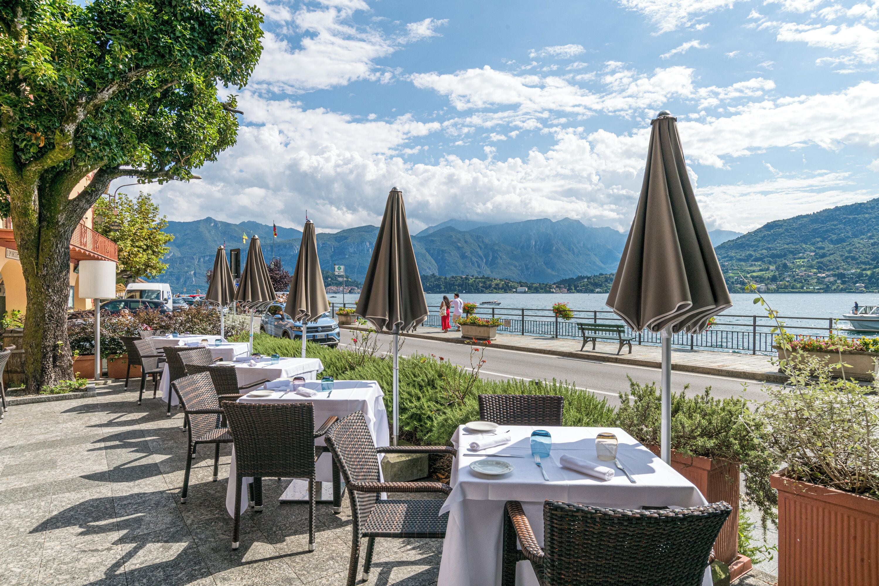 Ein Essbereich im Freien mit Korbstühlen und Tischen, Sonnenschirmen und Blick auf einen See und Berge.