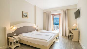A hotel room with two beds, a bedside table with a lamp, a TV mounted on the wall, and a window with curtains.