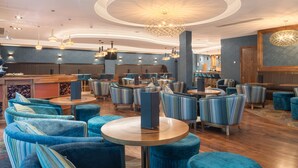A modern hotel lobby with round tables, blue upholstered chairs, and a central chandelier.