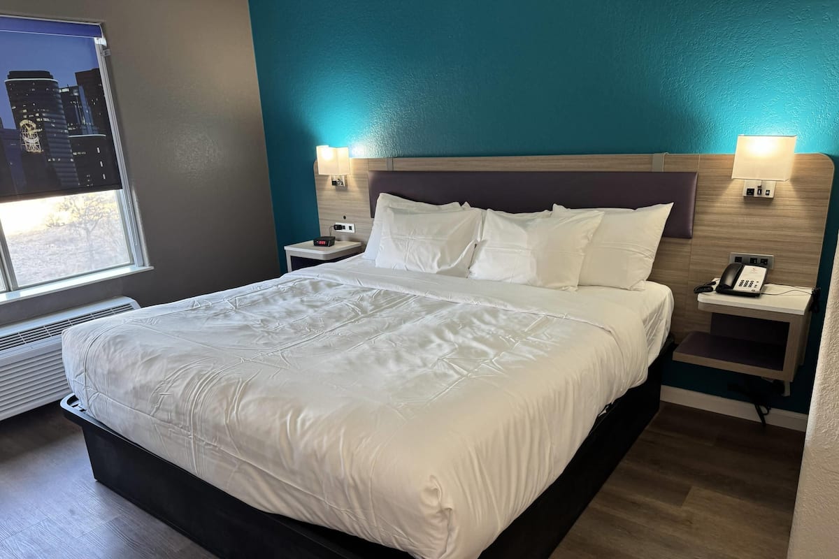 A hotel room with a large bed, a nightstand with a phone, a wall-mounted lamp, and a window with a city view.