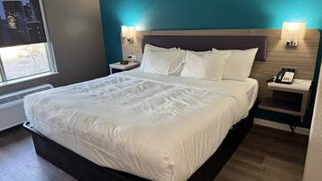 A hotel room with a large bed, a nightstand with a phone, a wall-mounted lamp, and a window with a city view.
