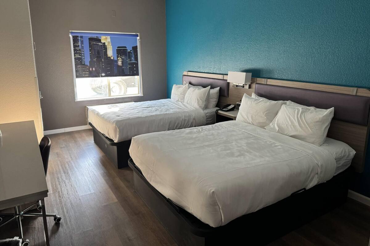 A hotel room with two beds, a desk, a chair, a window with a cityscape view, and a teal accent wall.