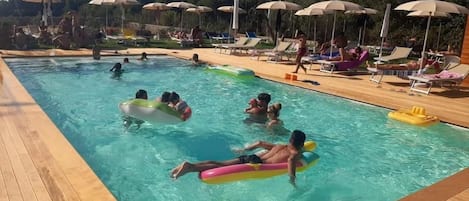 Una piscina con gente usando flotadores, rodeada de sillas de descanso y sombrillas.