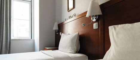 A hotel room with two beds, a nightstand, a lamp, a window with curtains, and a wooden headboard.
