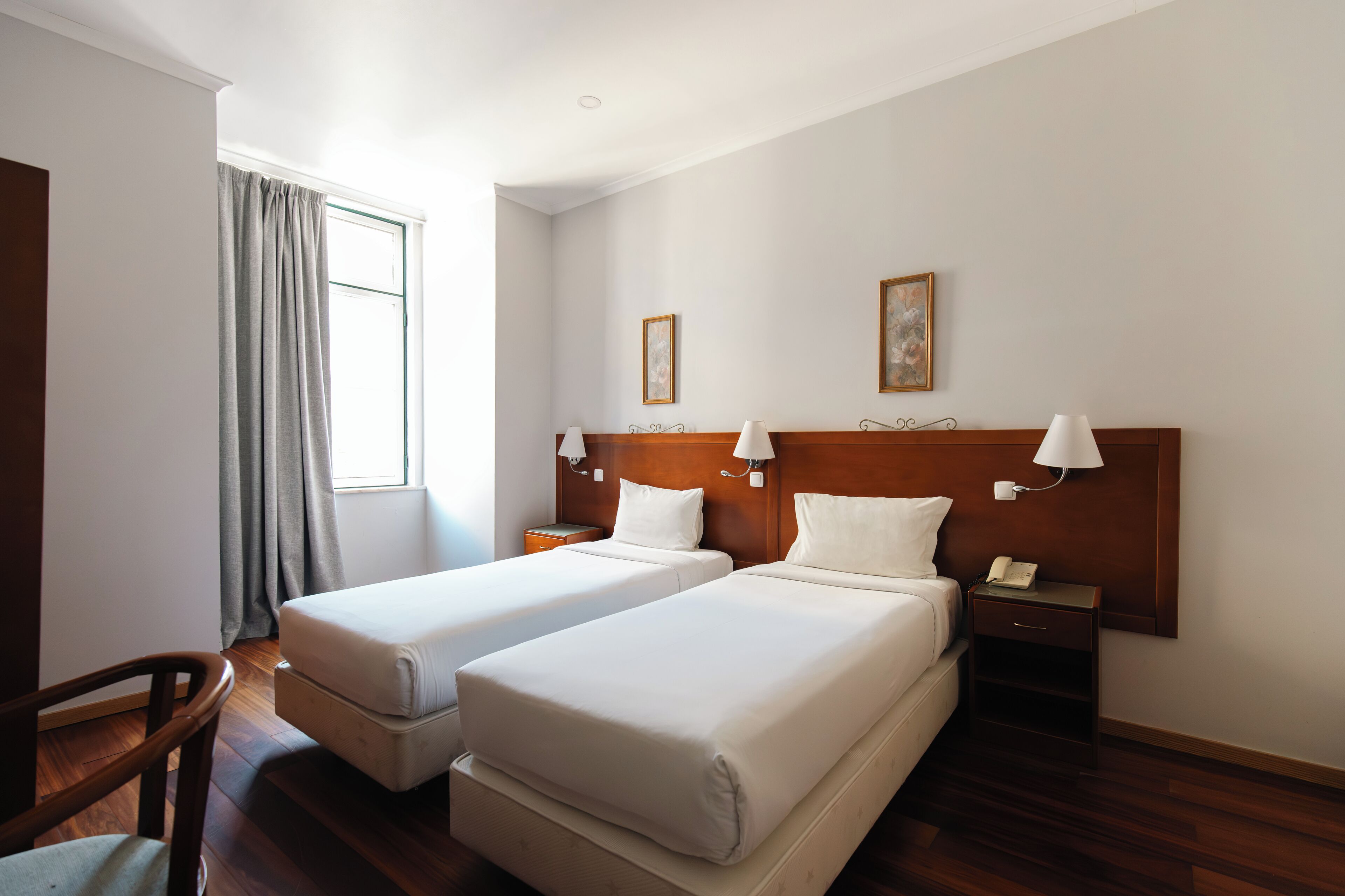 A hotel room with two beds, wooden headboards, bedside tables with lamps, a chair, and a window with curtains.
