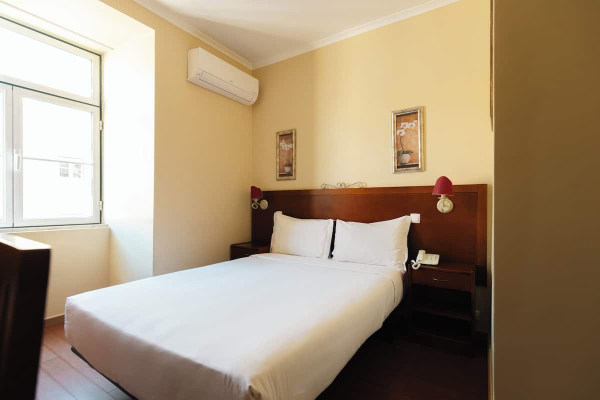 A hotel room with a large bed, a bedside table with a lamp, a clock, and a phone.