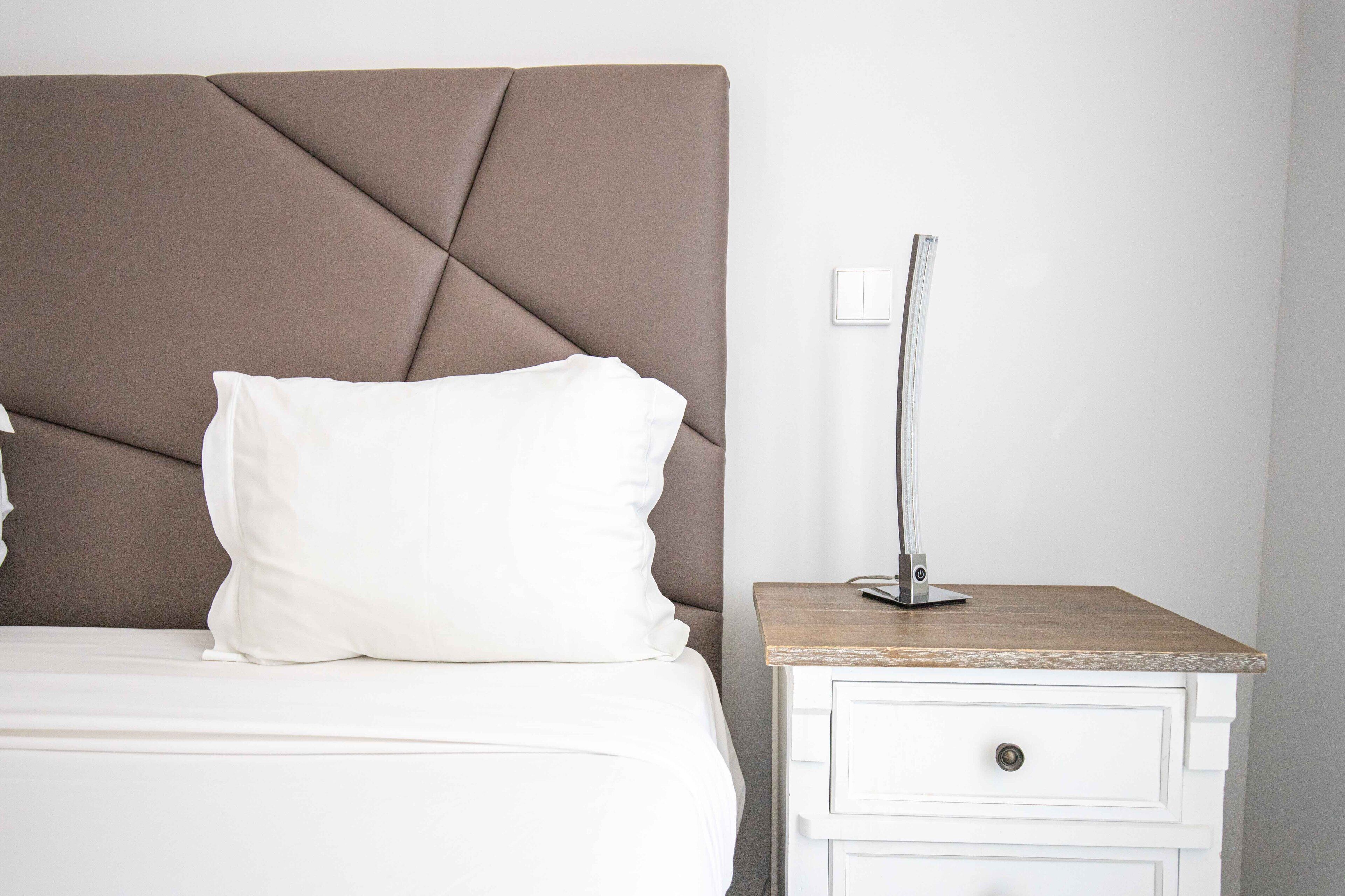 A neatly made bed with a headboard, a bedside lamp, and a small white nightstand with a drawer.