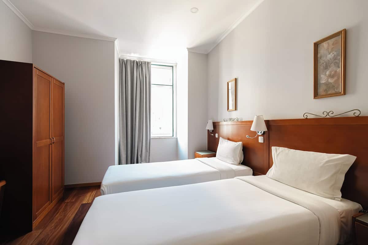 A hotel room with two single beds, a wooden wardrobe, a nightstand with a lamp, and a window with curtains.