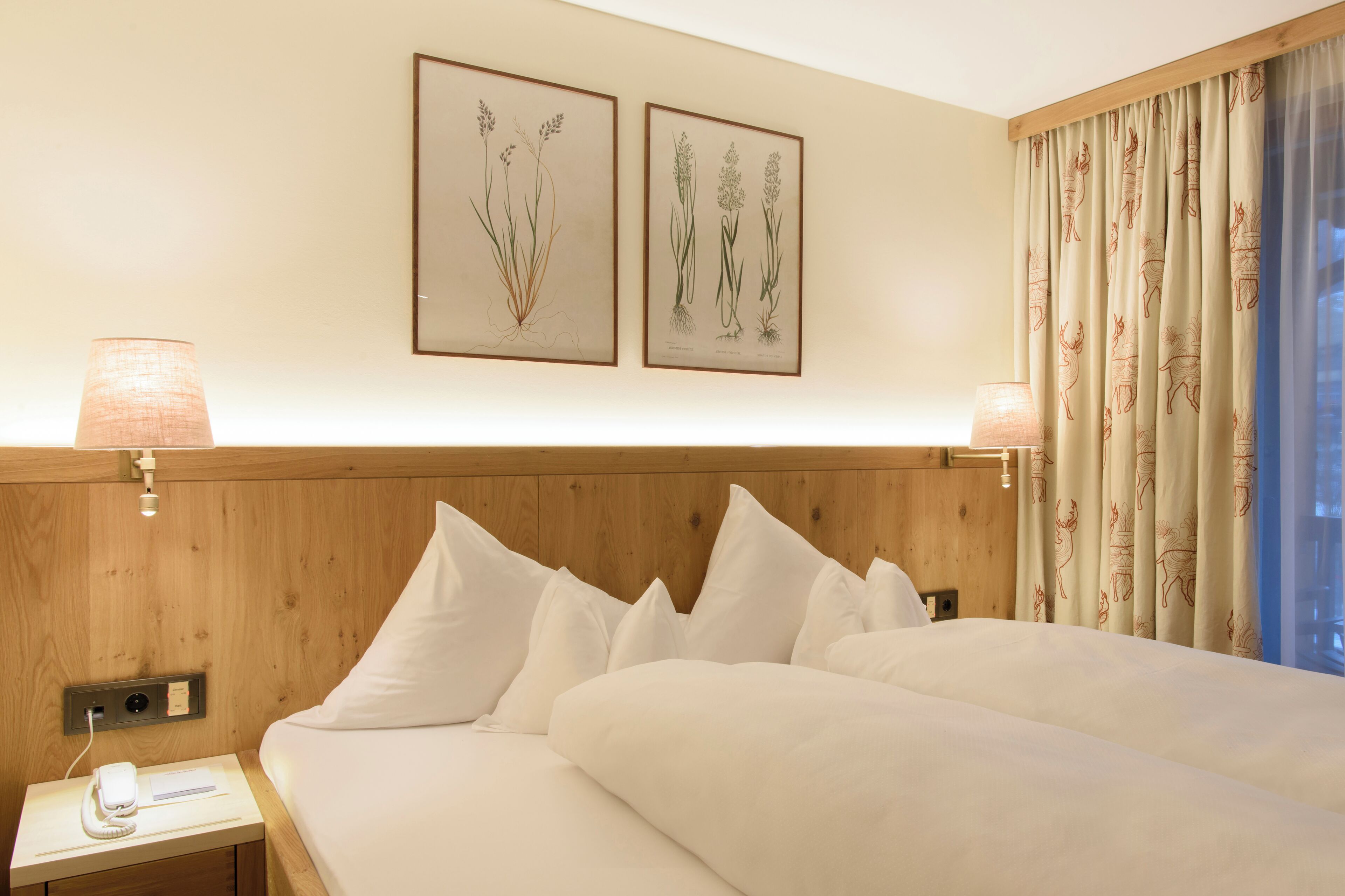 A hotel room with a wooden headboard, white bedding, two framed botanical prints on the wall, and a window with patterned curtains.