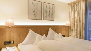 A hotel room with a wooden headboard, white bedding, two framed botanical prints on the wall, and a window with patterned curtains.