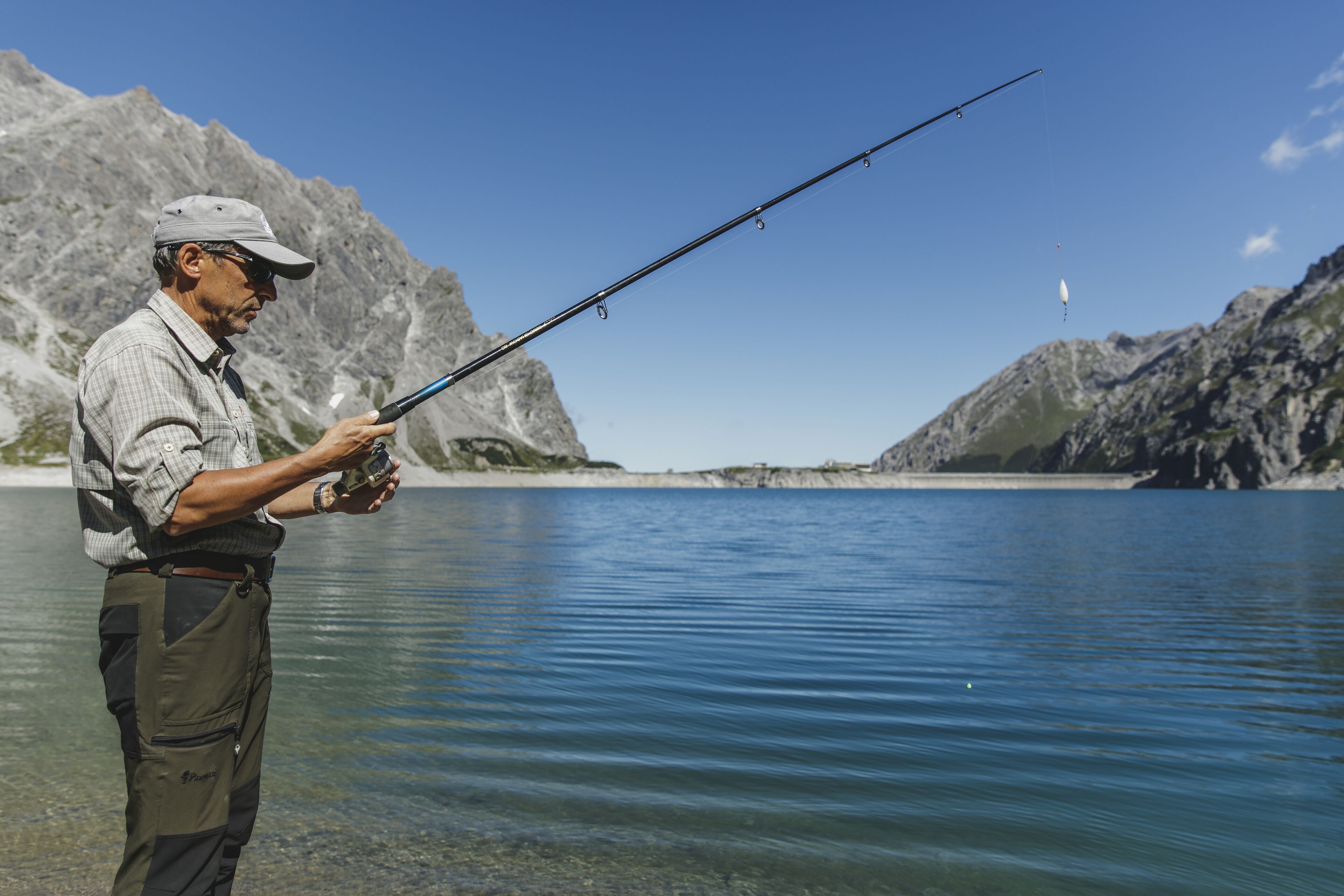 Une personne est en train de pêcher au bord d’un lac paisible, avec des montagnes en arrière-plan.