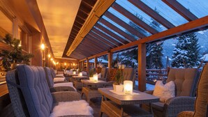 An outdoor seating area with wicker furniture and tables, under a wooden pergola with a view of snow-covered trees. - Montafoner Hof (Tschagguns)