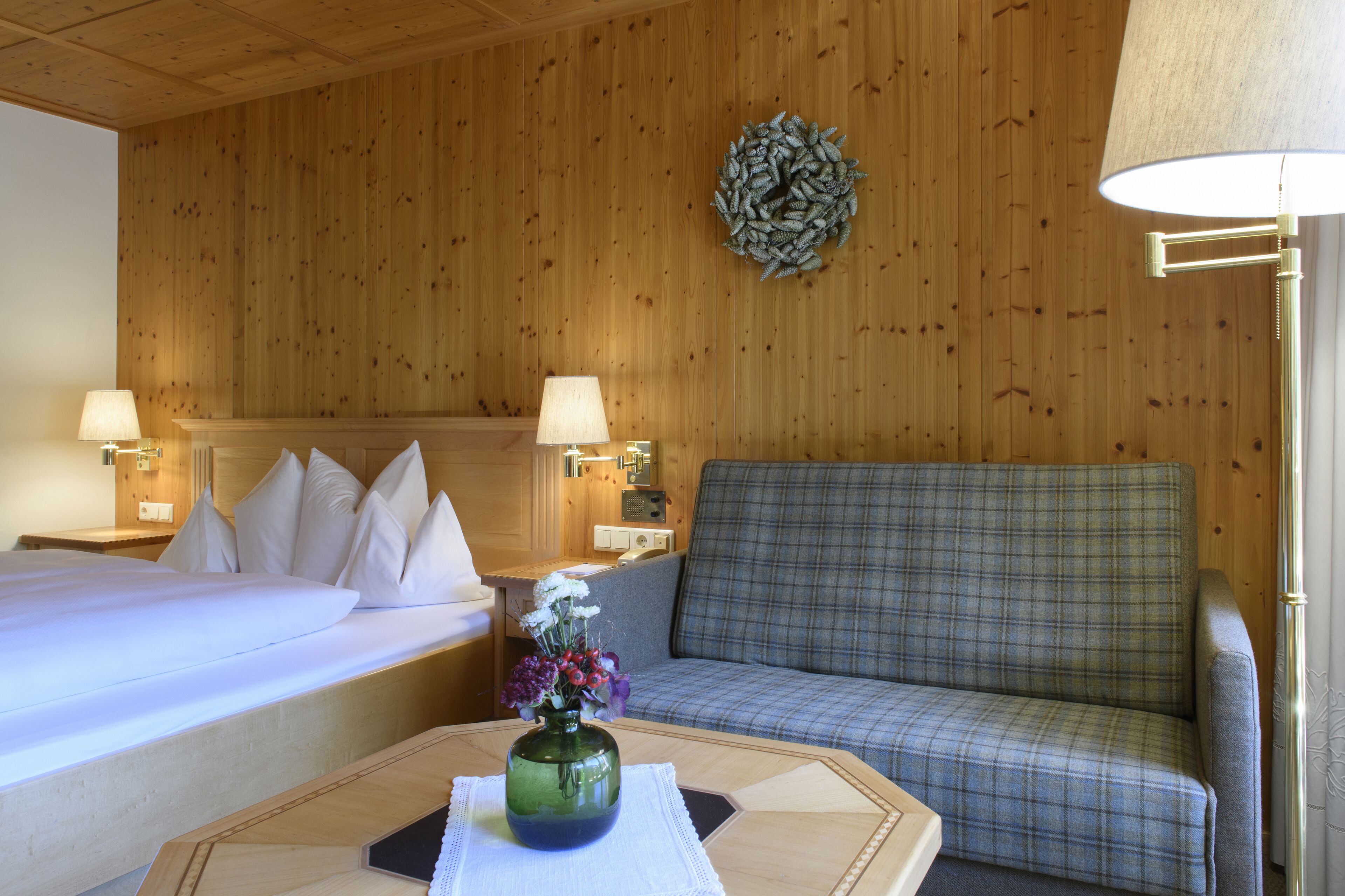 A hotel room with a bed, a sofa, a table with a vase of flowers, and two wall-mounted lamps.