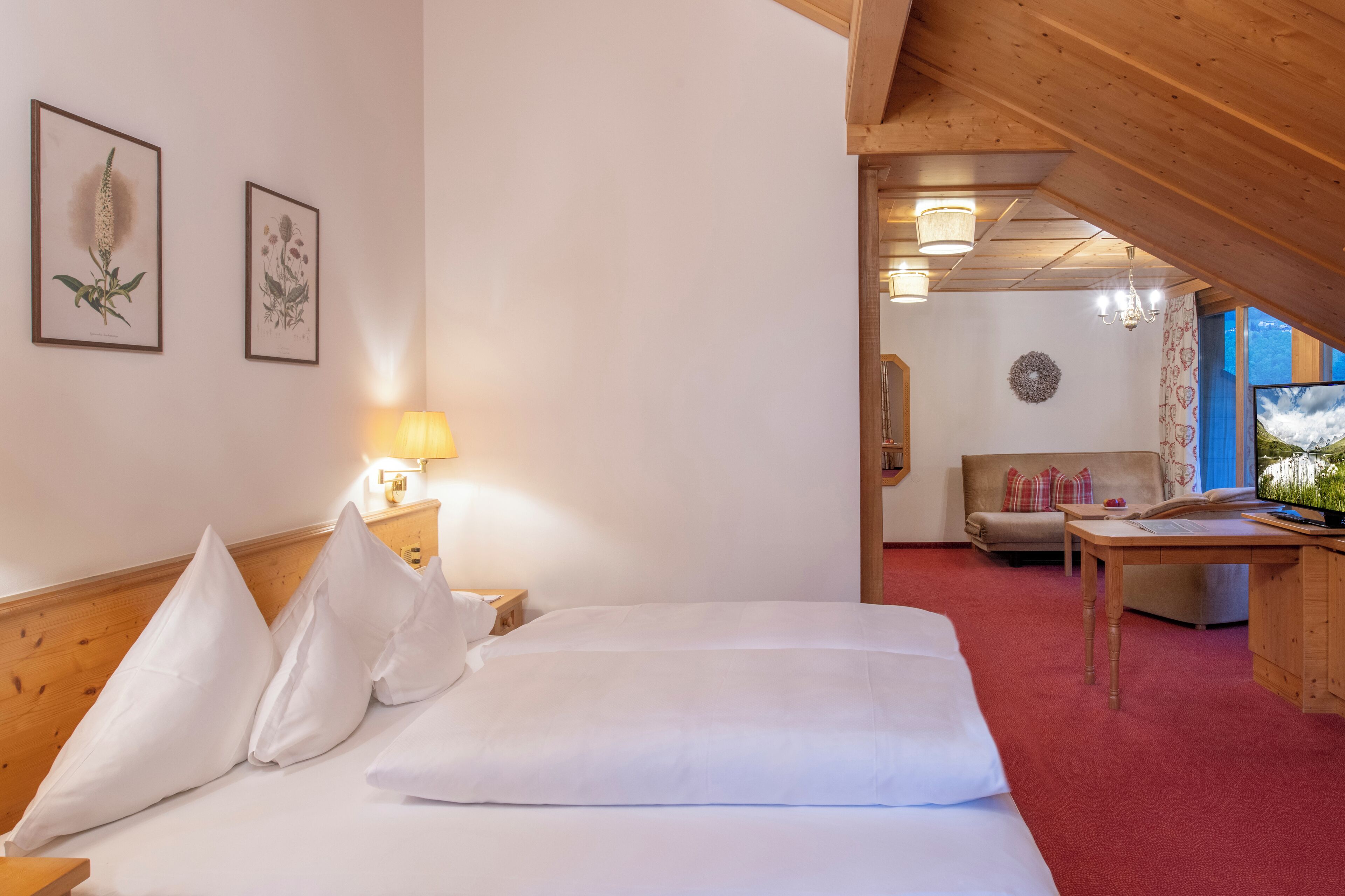 Ein Hotelzimmer mit Holzboden, einem Bett mit weißer Bettwäsche, einem Nachttisch aus Holz mit einer Lampe und gerahmten Pflanzenmotiven an der Wand.