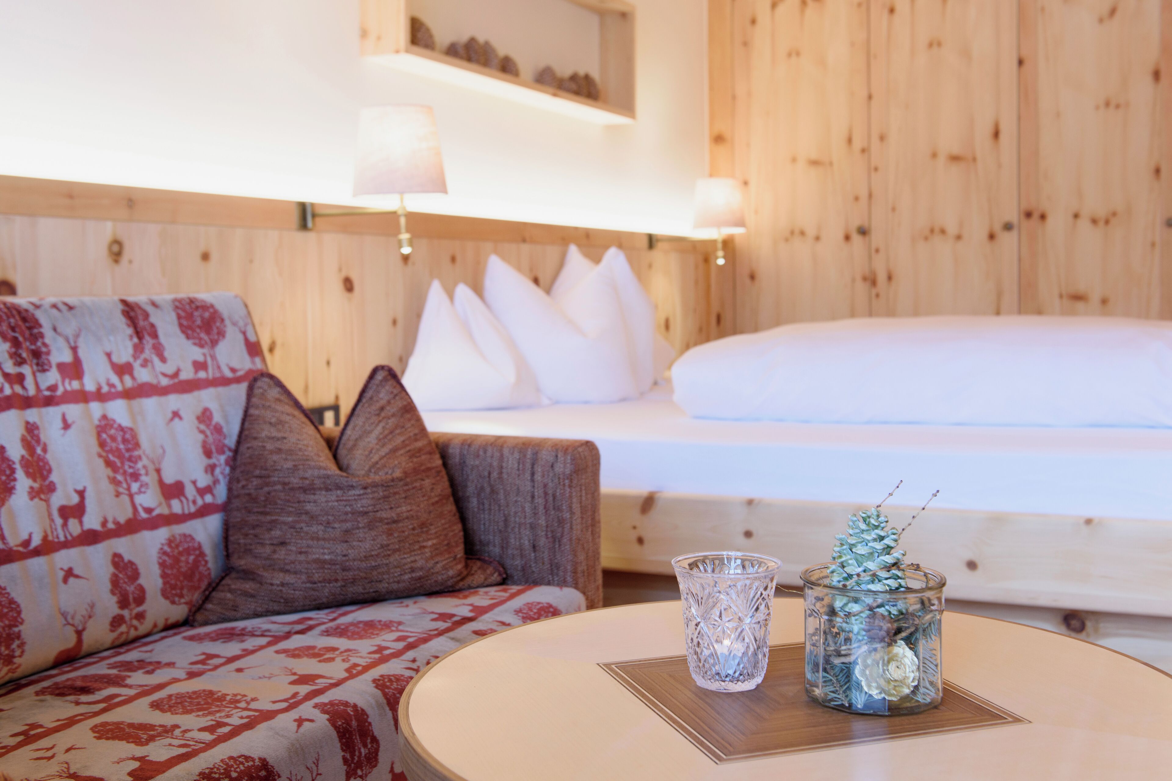 A cozy room with a wooden wall, a bed with white bedding, a patterned sofa, and a small table with a glass vase and pinecone decoration.