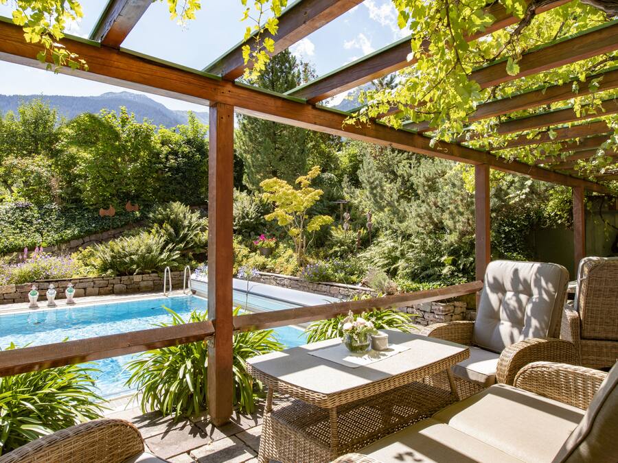 A covered patio with a pool, wicker furniture, and a view of a lush garden and mountains.