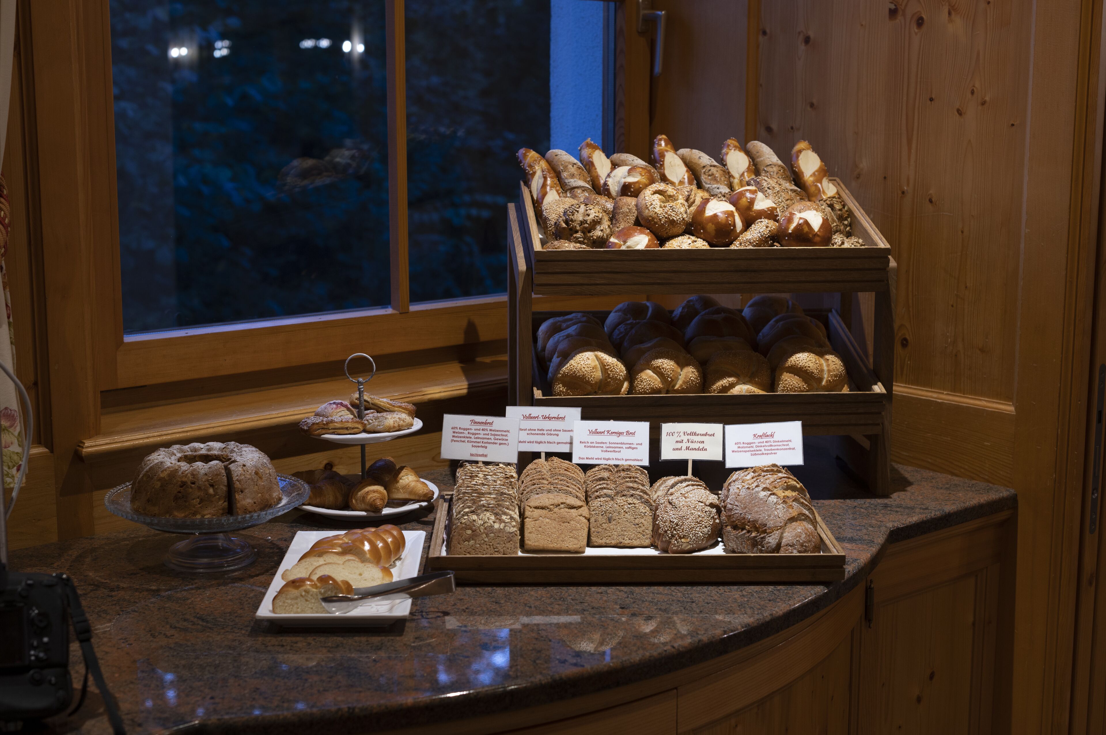 Une vitrine de boulangerie présentant diverses sortes de pains et de pâtisseries, dont un gâteau sur un présentoir, le tout disposé sur un comptoir près d’une fenêtre.