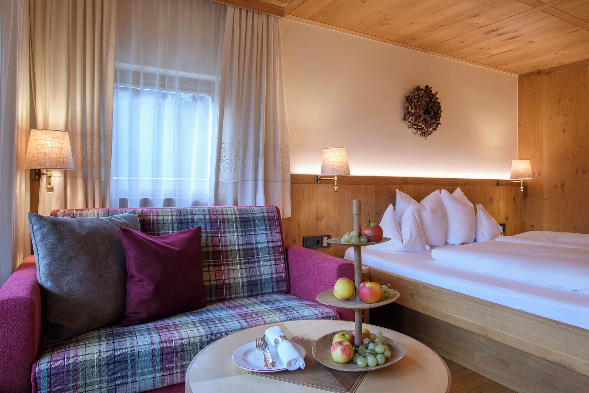 A hotel room with a plaid sofa, a bed, a small round table with a fruit and napkin set, and two wall-mounted lamps.