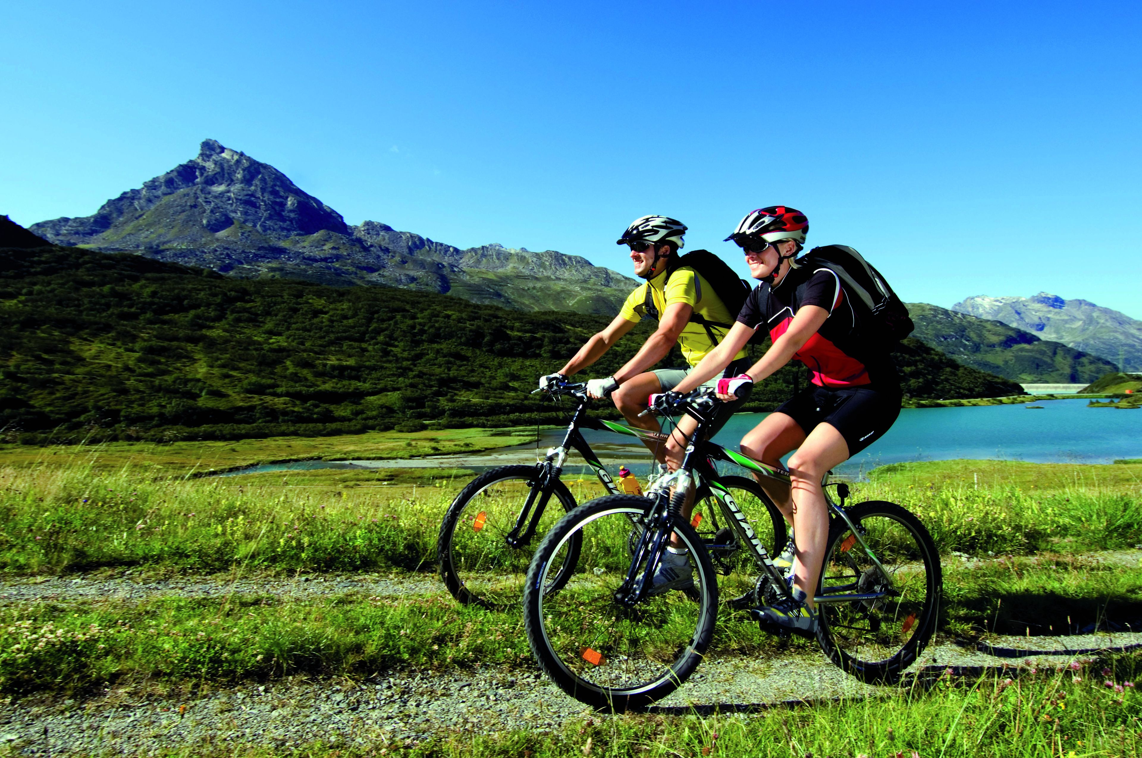 Deux cyclistes sur un sentier de VTT, avec un lac et des montagnes en arrière-plan.
