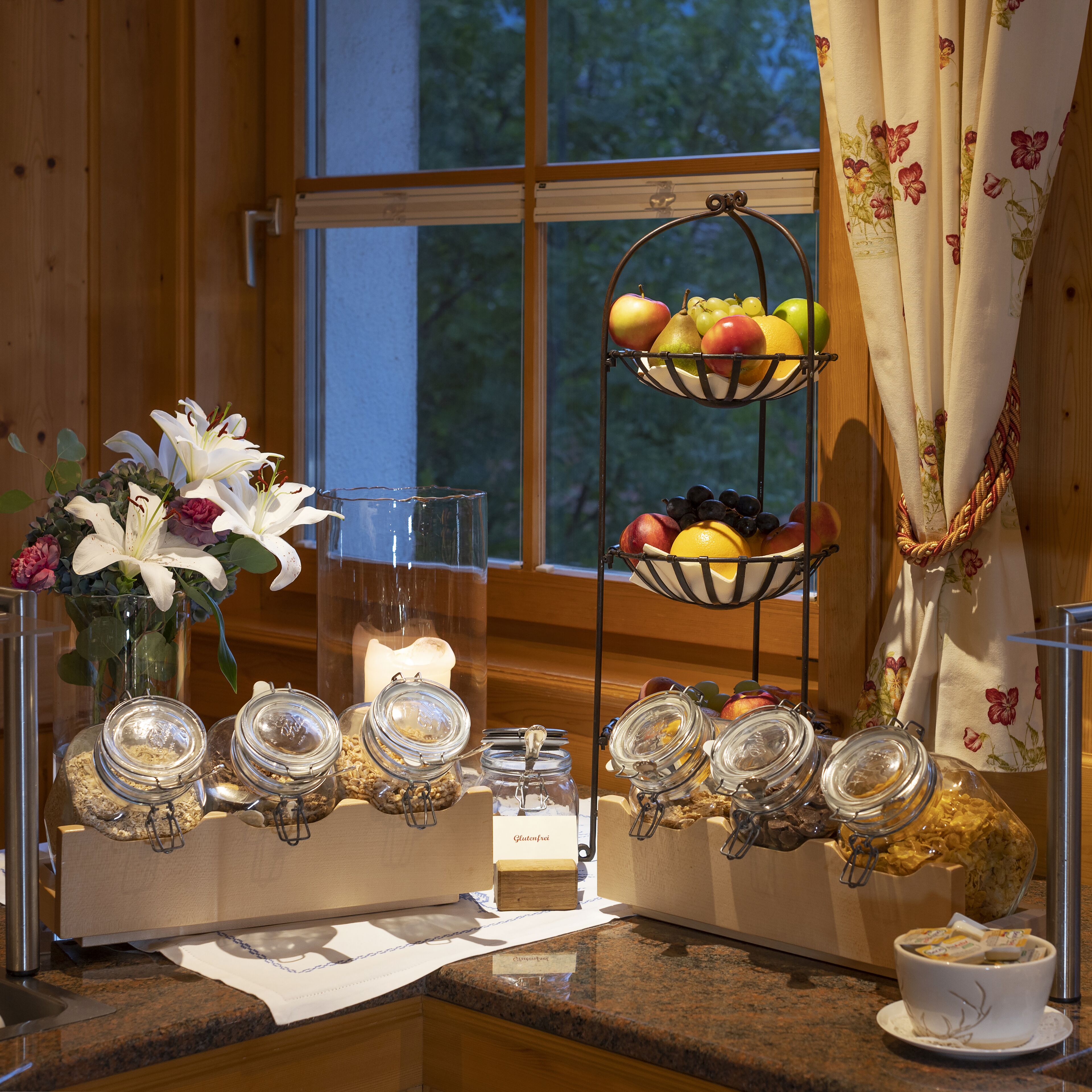 A breakfast setup with a fruit basket, a candle, and jars of food on a wooden counter near a window.