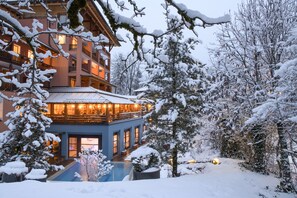 Un hotel de varios pisos con zona de piscina, rodeado de árboles nevados y un cielo despejado.