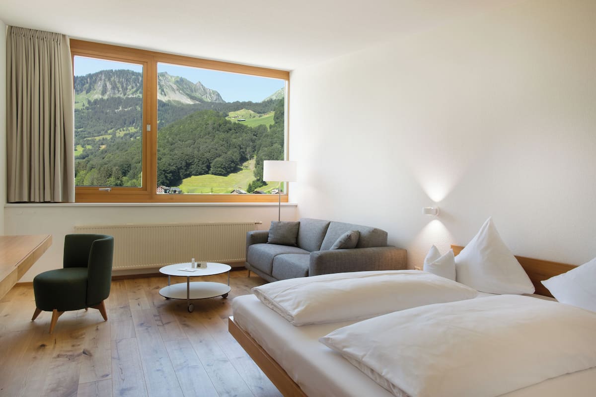 A hotel room with a bed, a sofa, a chair, a small table, and a view of a mountainous landscape through a large window.