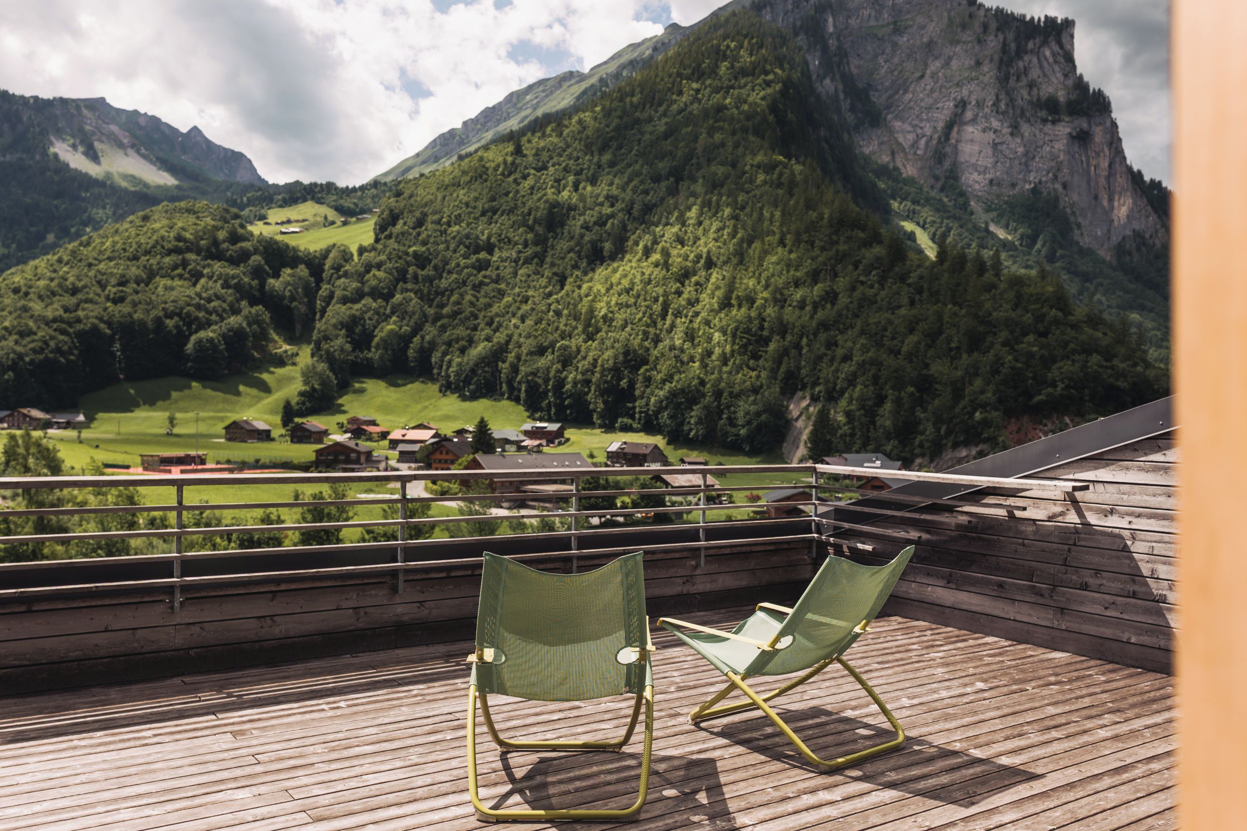 A balcony with two green chairs overlooking a mountainous landscape with a village below.