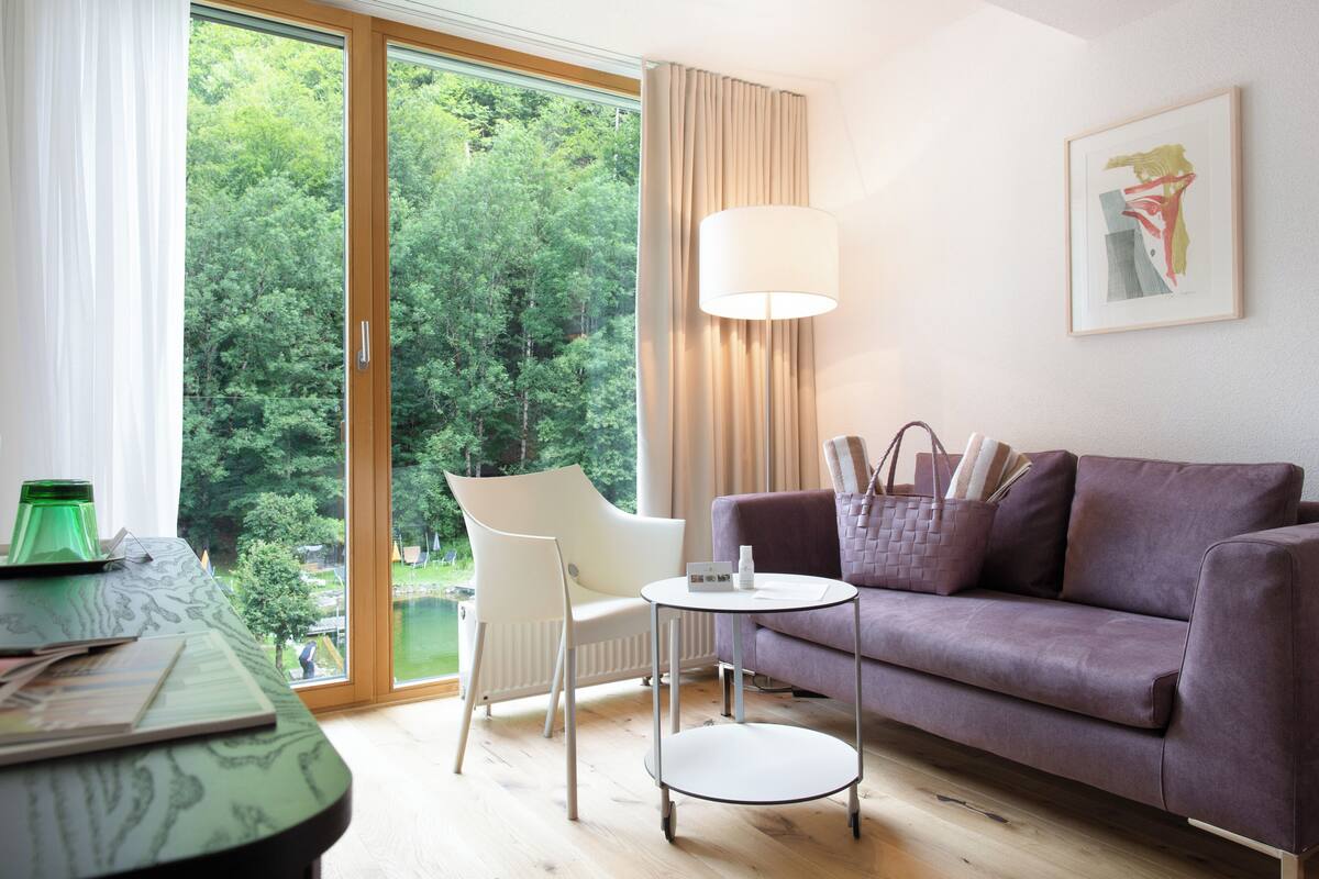 A modern living room with a sofa, a table, and a chair. There is a window with a view of trees and a lamp.