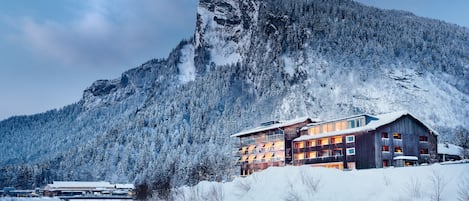 Una cabaña de montaña con un río frente a ella, rodeada de árboles cubiertos de nieve y una montaña rocosa.