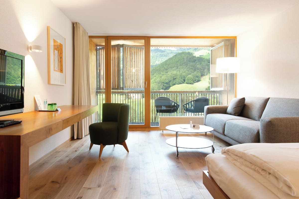 A hotel room with a wooden desk, a green armchair, a grey sofa, a round coffee table, and a bed.