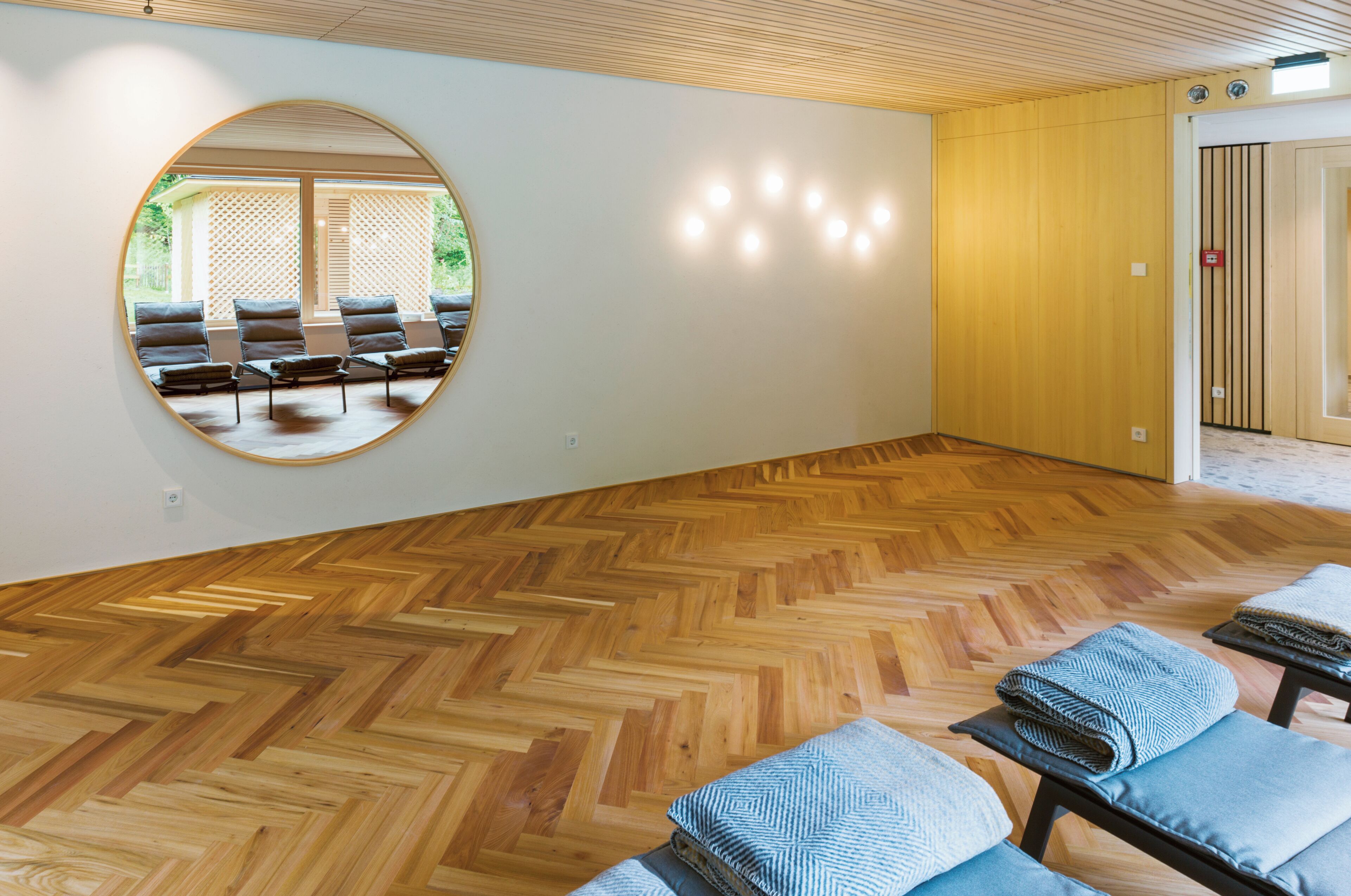 A room with a herringbone wooden floor, a round mirror on the wall, and a view of an outdoor seating area through the mirror.