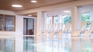 A spacious indoor pool area with clear glass walls, a wooden ceiling, and several lounge chairs arranged for relaxation. - Hotel Krone in Au (Au)