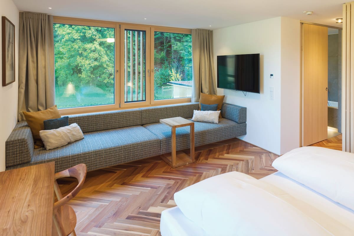 A modern hotel room with a large window, a sofa, a bed, and a television.