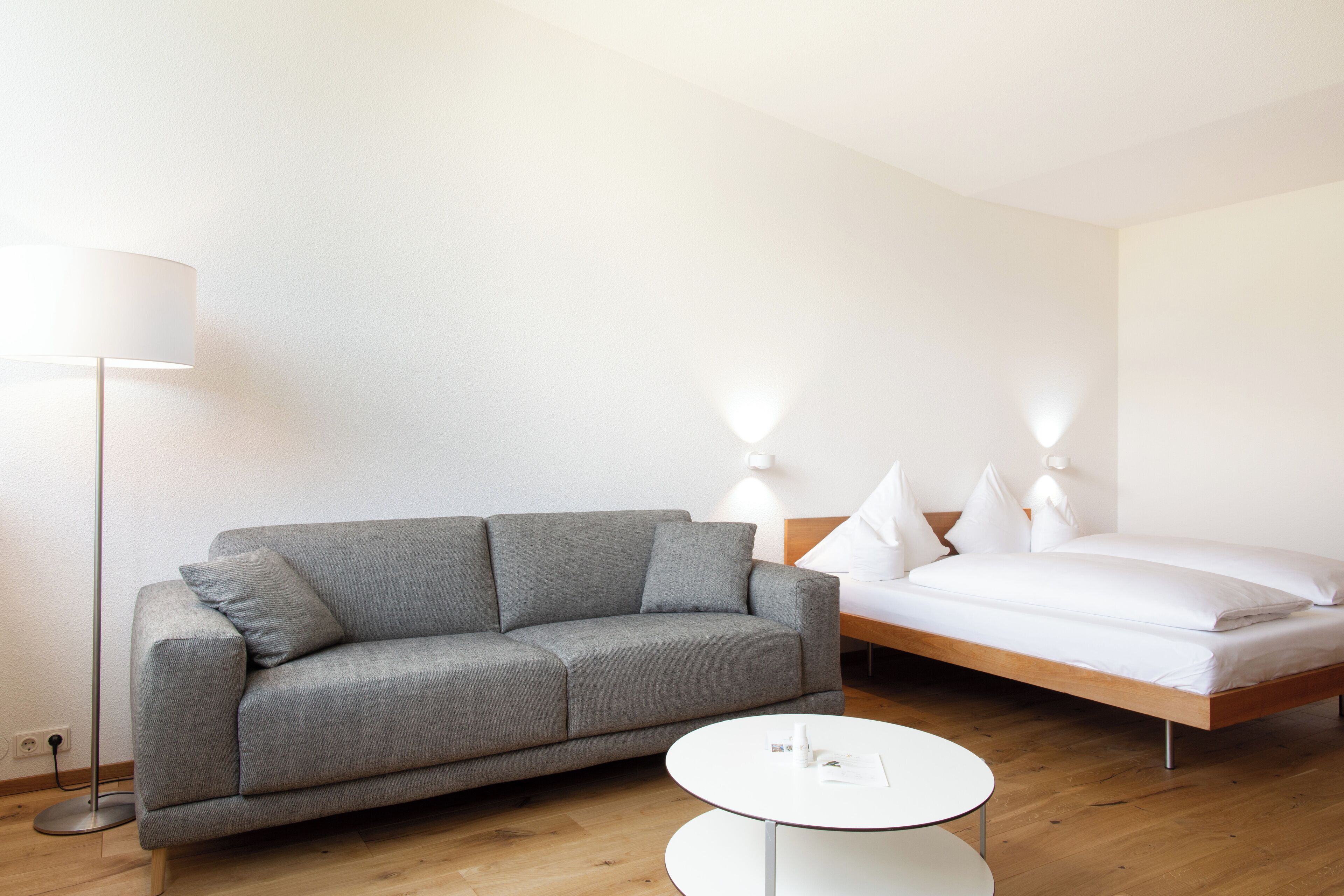 Ein modernes Hotelzimmer mit grauem Sofa, einem hölzernen Bett, einem weißen Nachttisch und einer Stehlampe.