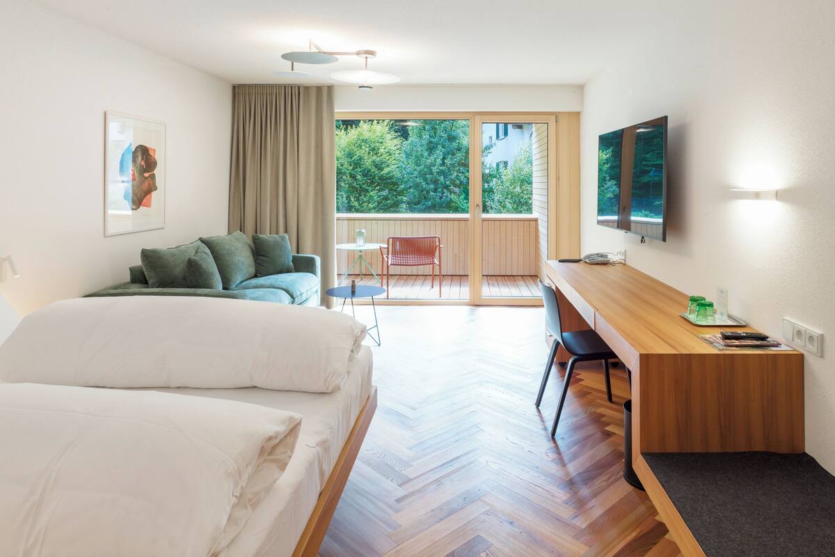 A hotel room with a bed, a desk, a chair, a TV, and a balcony with a view of trees.