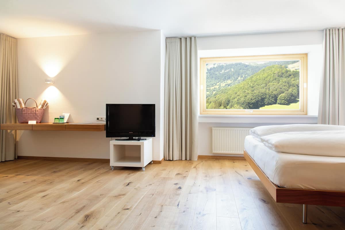 A hotel room with a wooden floor, a bed, a TV on a stand, a window with a view of a forest, and a shelf with a basket and some items.