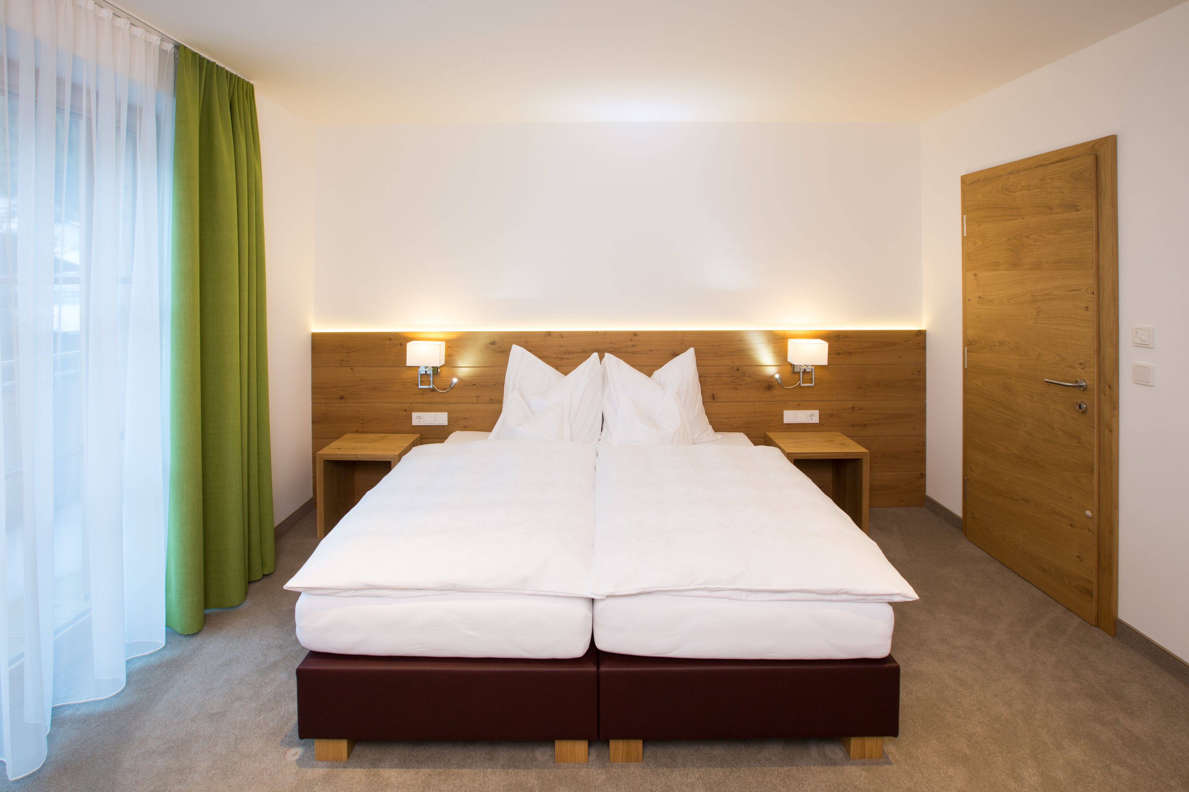 Cama doble con cabecera de madera, dos mesitas de noche con lámparas, una puerta de madera y cortinas verdes.