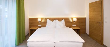 Cama doble con cabecera de madera, dos mesitas de noche con lámparas, una puerta de madera y cortinas verdes.