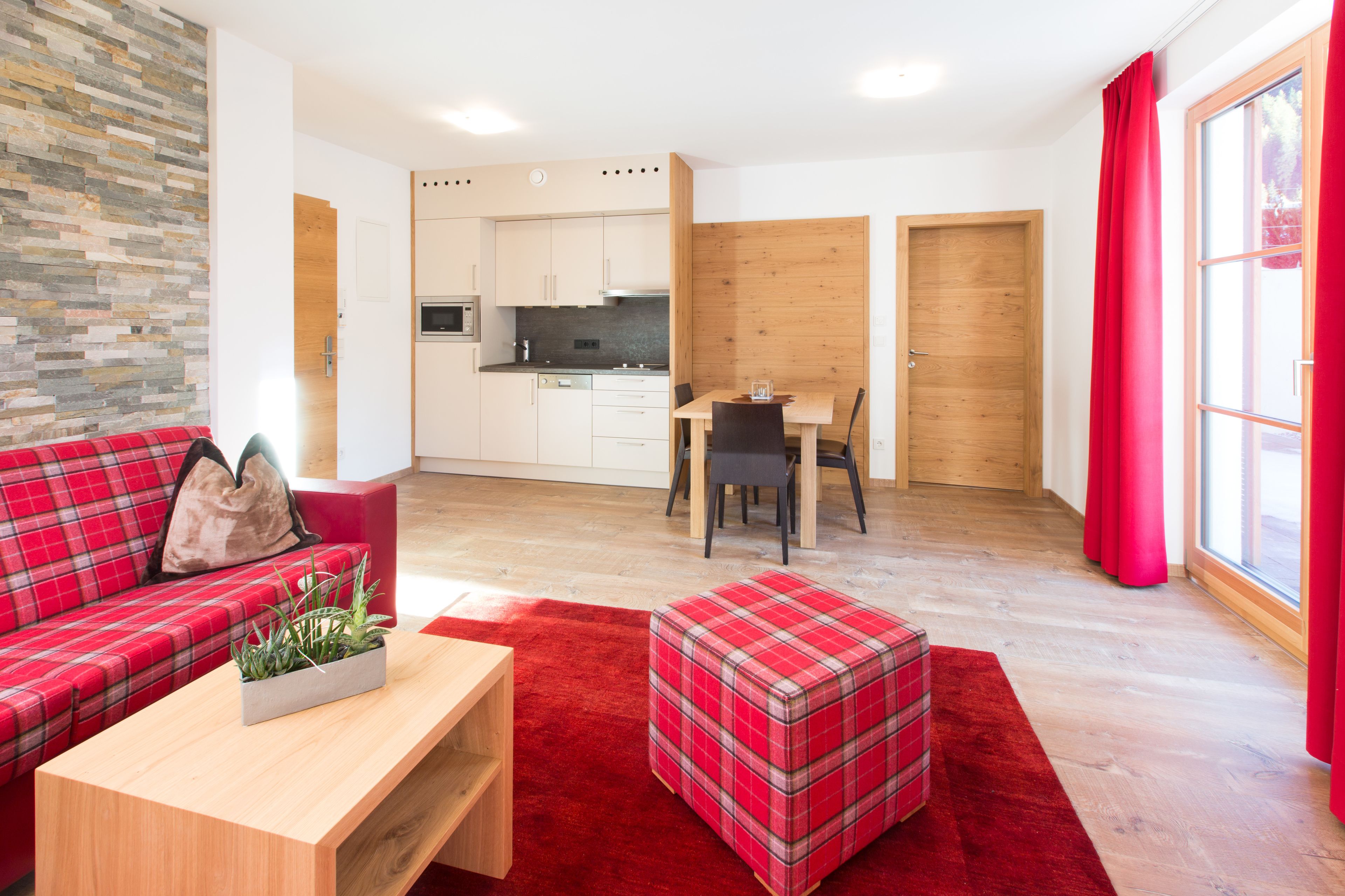 Una sala de estar moderna con un sofá de cuadros rojos, una mesa de centro de madera con una planta y un reposapiés a cuadros. Se ve una cocina con armarios blancos y un comedor con una mesa y sillas al fondo.