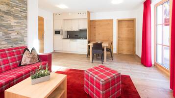 Una sala de estar moderna con un sofá de cuadros rojos, una mesa de centro de madera con una planta y un reposapiés a cuadros. Se ve una cocina con armarios blancos y un comedor con una mesa y sillas al fondo.