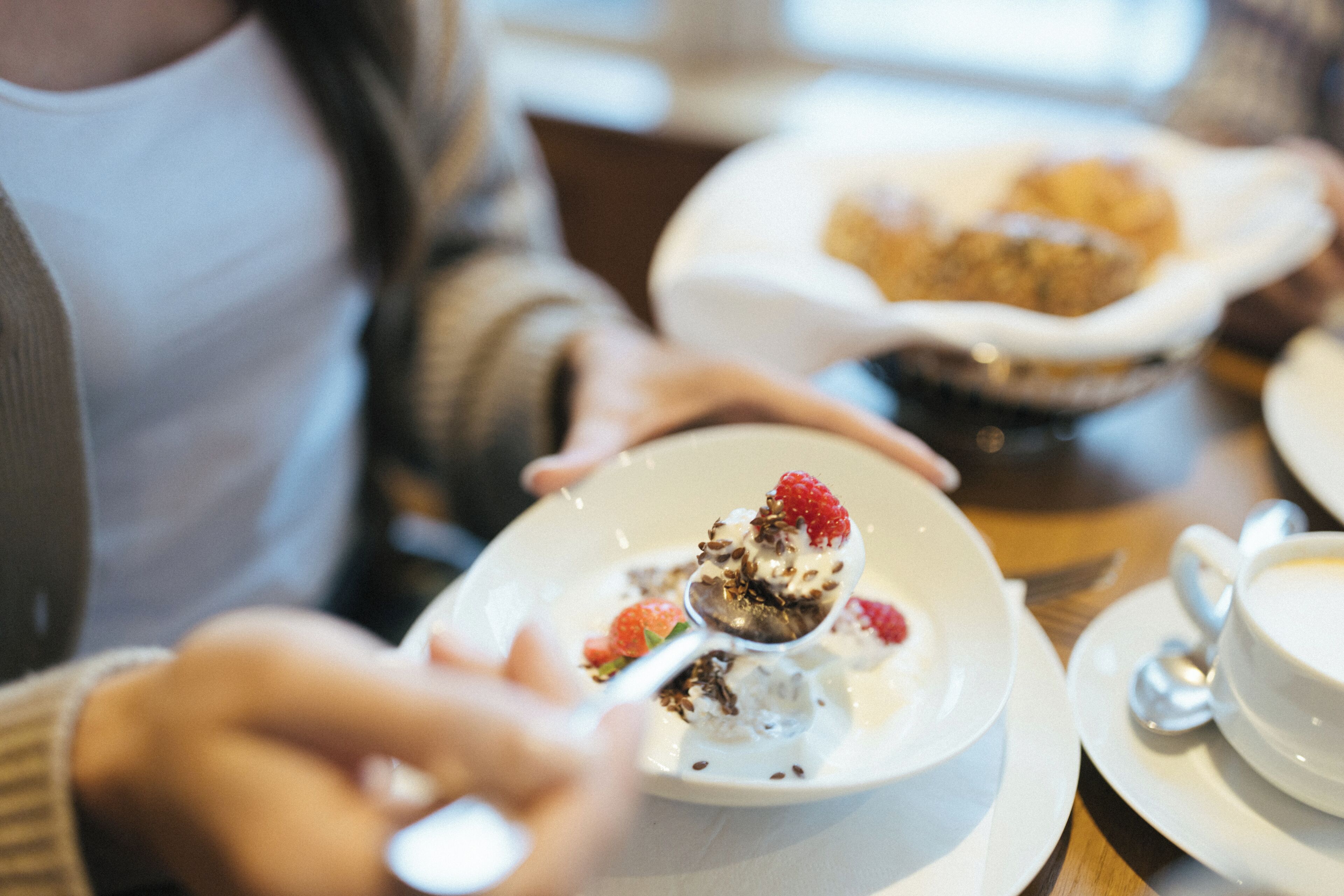Una mesa de comedor con un plato blanco que contiene un postre cubierto con frambuesas, una cuchara y una taza blanca sobre un platito.