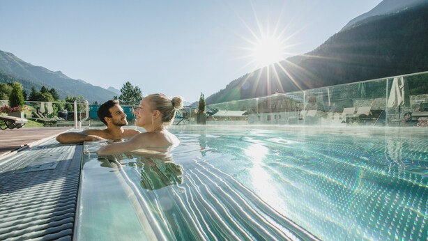 Una pareja descansando en una piscina con fondo de cristal y montañas al fondo.