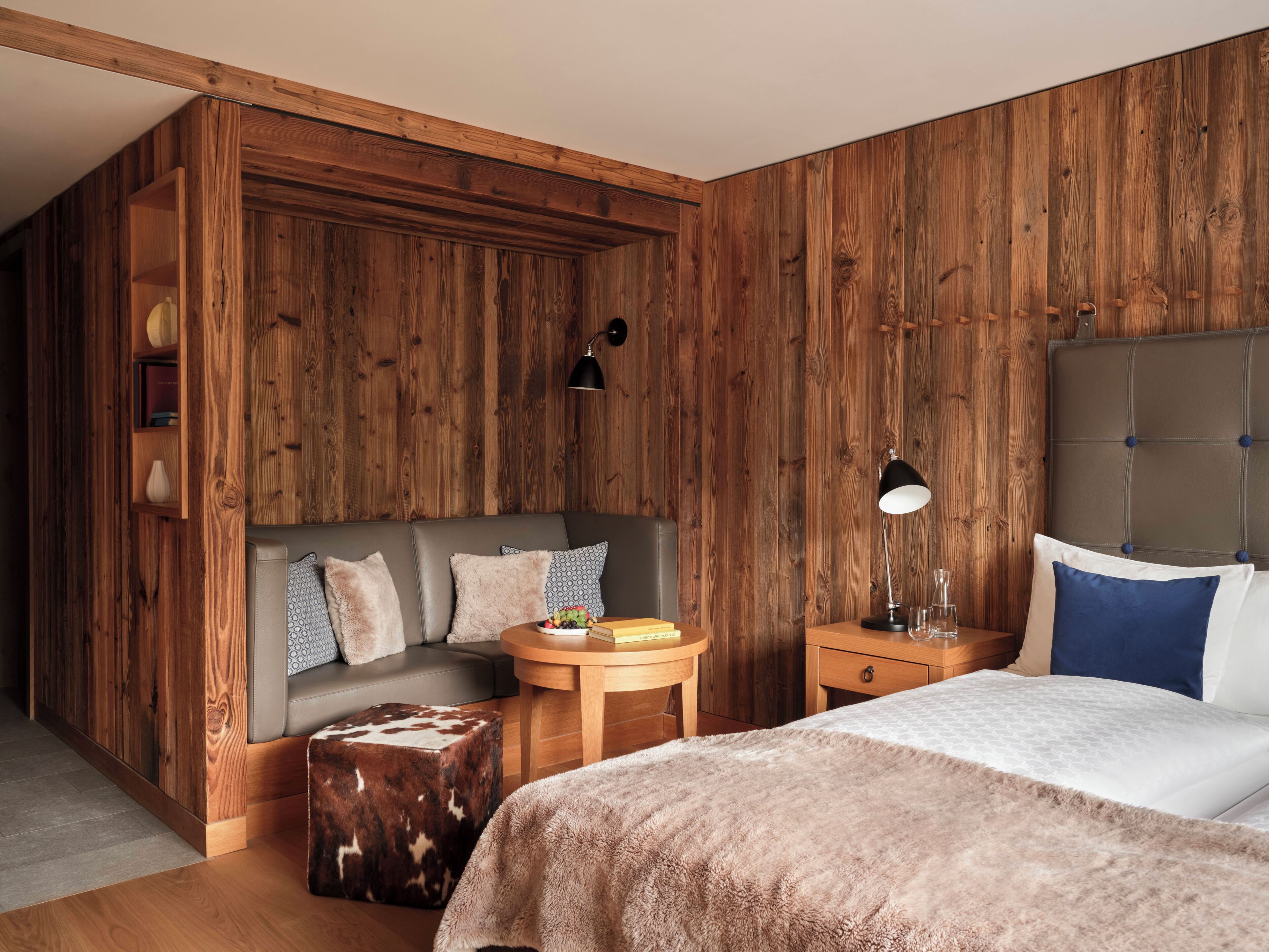 A bedroom with a wooden panel wall, a sofa, a round wooden table, a bedside table, and a bed with a tufted headboard.