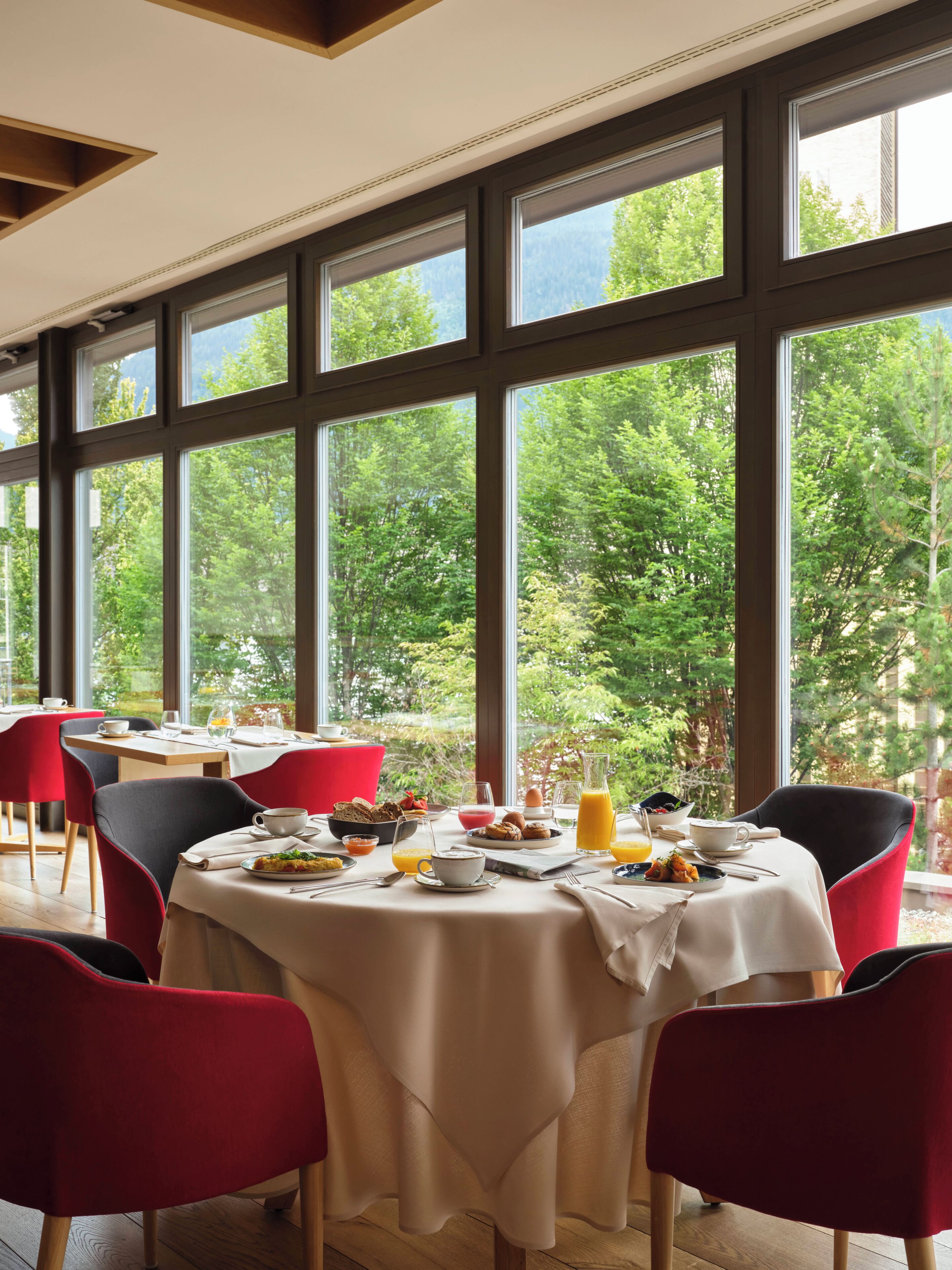Ein Essbereich mit Tisch, gedeckt für das Frühstück, großen Fenstern mit Blick auf einen Wald und roten Stühlen.